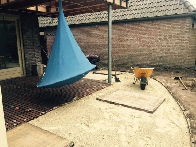 Blauwe hangstoel boven een houten vlonder, met een kruiwagen ernaast